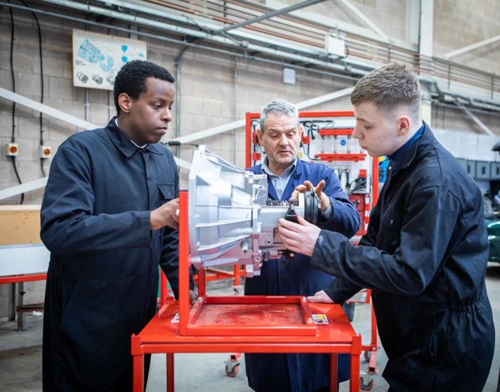 Automotive instructor guiding two students in overalls as they work on a mechanical component in a workshop. Automotive instructor guiding two students in overalls as they work on a mechanical component in a workshop.